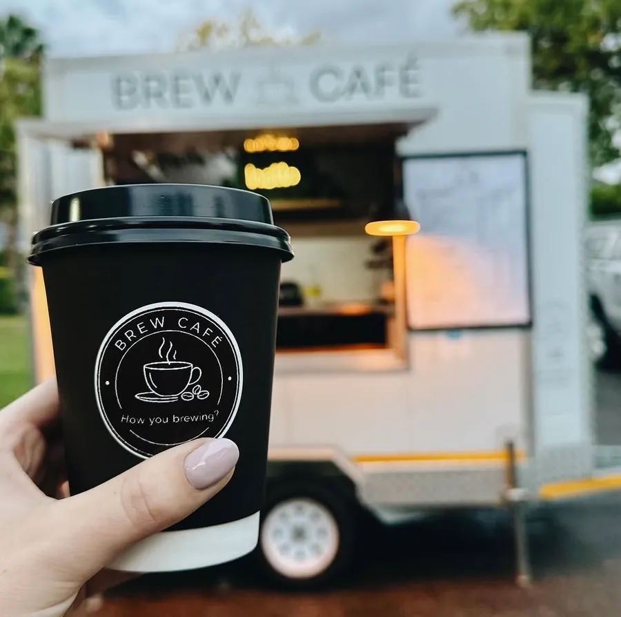 A hand holding a Brew Café branded coffee cup in front of the coffee trailer.