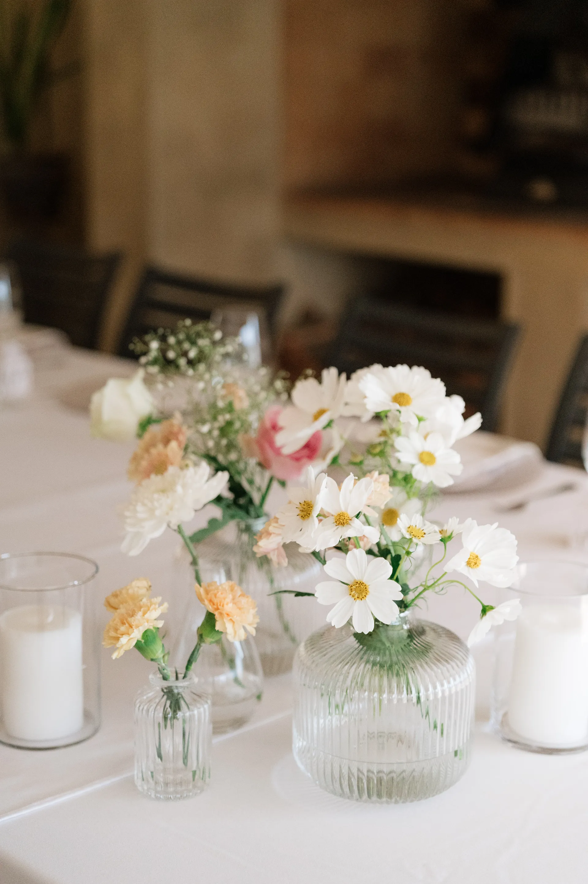 A blurred wedding reception venue with soft bokeh lighting and pastel florals in glass bud vases in the foreground.